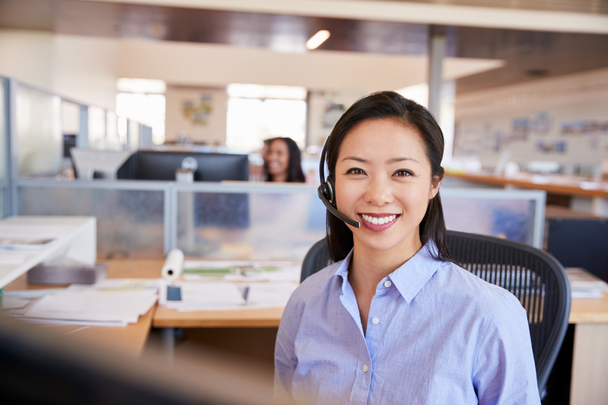 Female customer service representative wearing a microphone headset, sitting at a computer. Other customer service representatives are visible in the background.