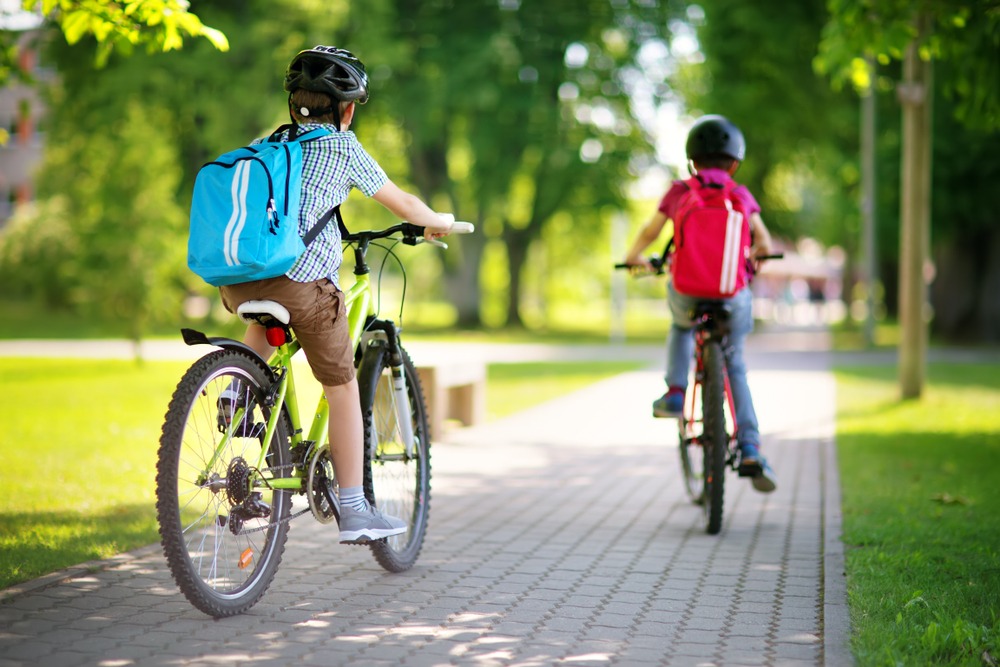 Two children riding bicycles through a park, on a sunny day, viewed from behind. The bike path is made of tidy pavers. Both children are wearing backpacks and helmets. Rockpoint Legal Funding is among the best lawsuit loan companies and provides cash advances for plaintiffs in bicycle accident lawsuits.